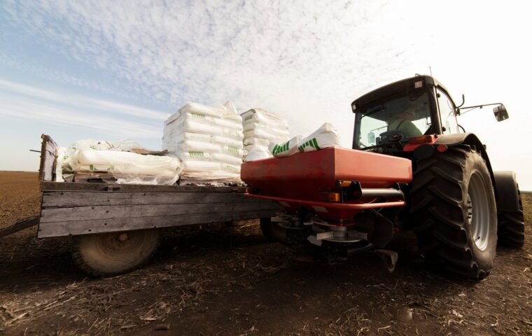 A tractor pulls a flatbed trailer stacked with bagged fertilizer and a spreader applicator across a California field