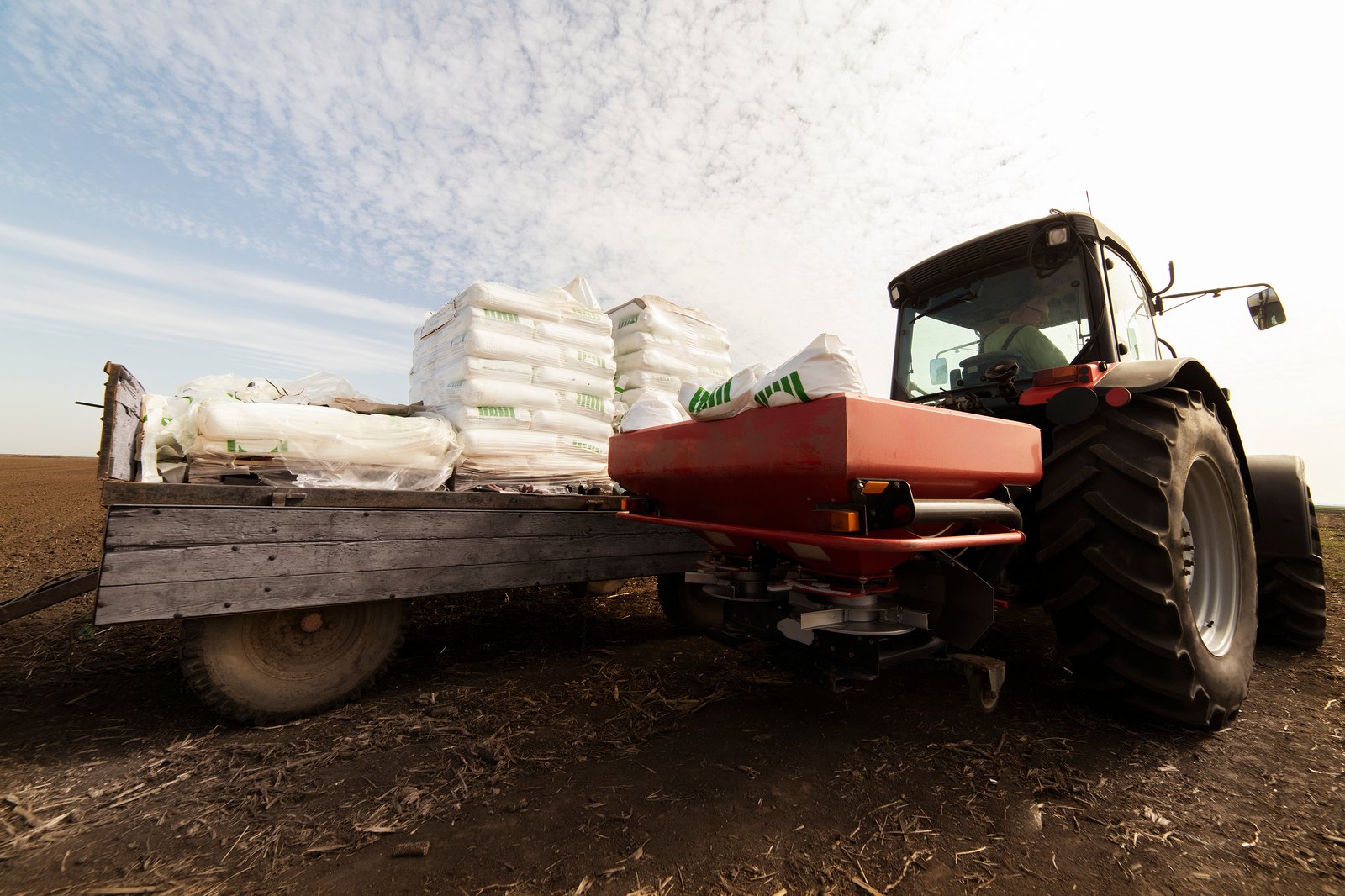 A tractor pulls a flatbed trailer stacked with bagged fertilizer and a spreader applicator across a California field