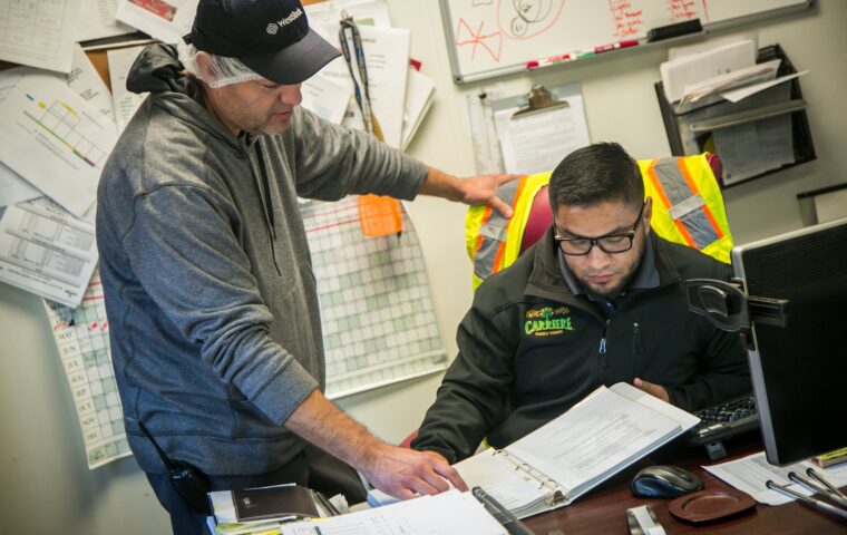 A farm supervisor and a worker in a high-visibility vest review a binder of safety documentation together at an office desk