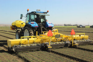 Tractor pulling a wide yellow implement across planted rows in a field, with a water tank mounted on the tractor.