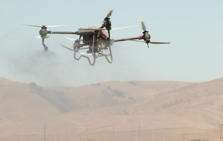 Agricultural spray drone flying low over a field with dry foothills in the background